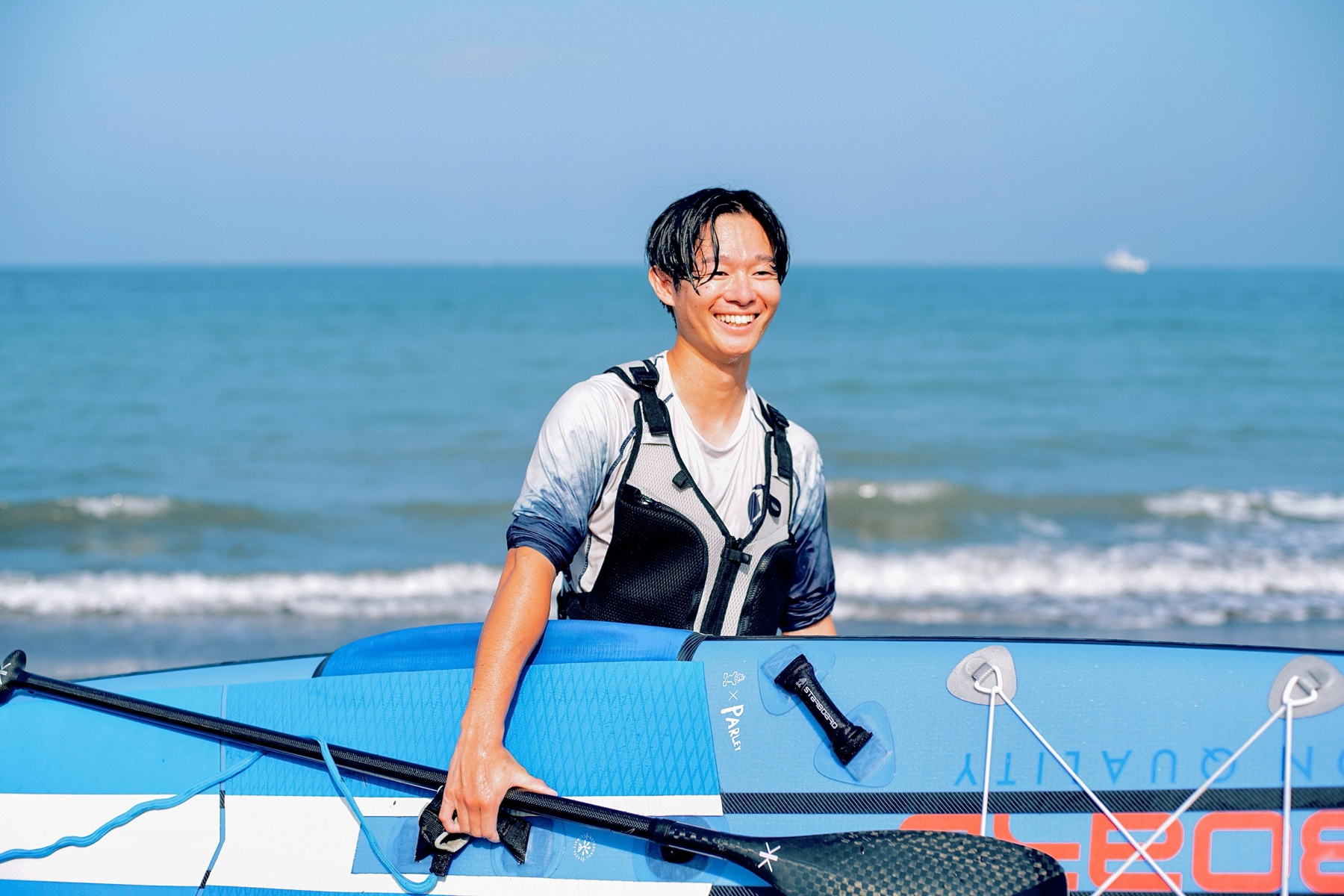 SUPボードを手に海辺で笑う濱中怜雄